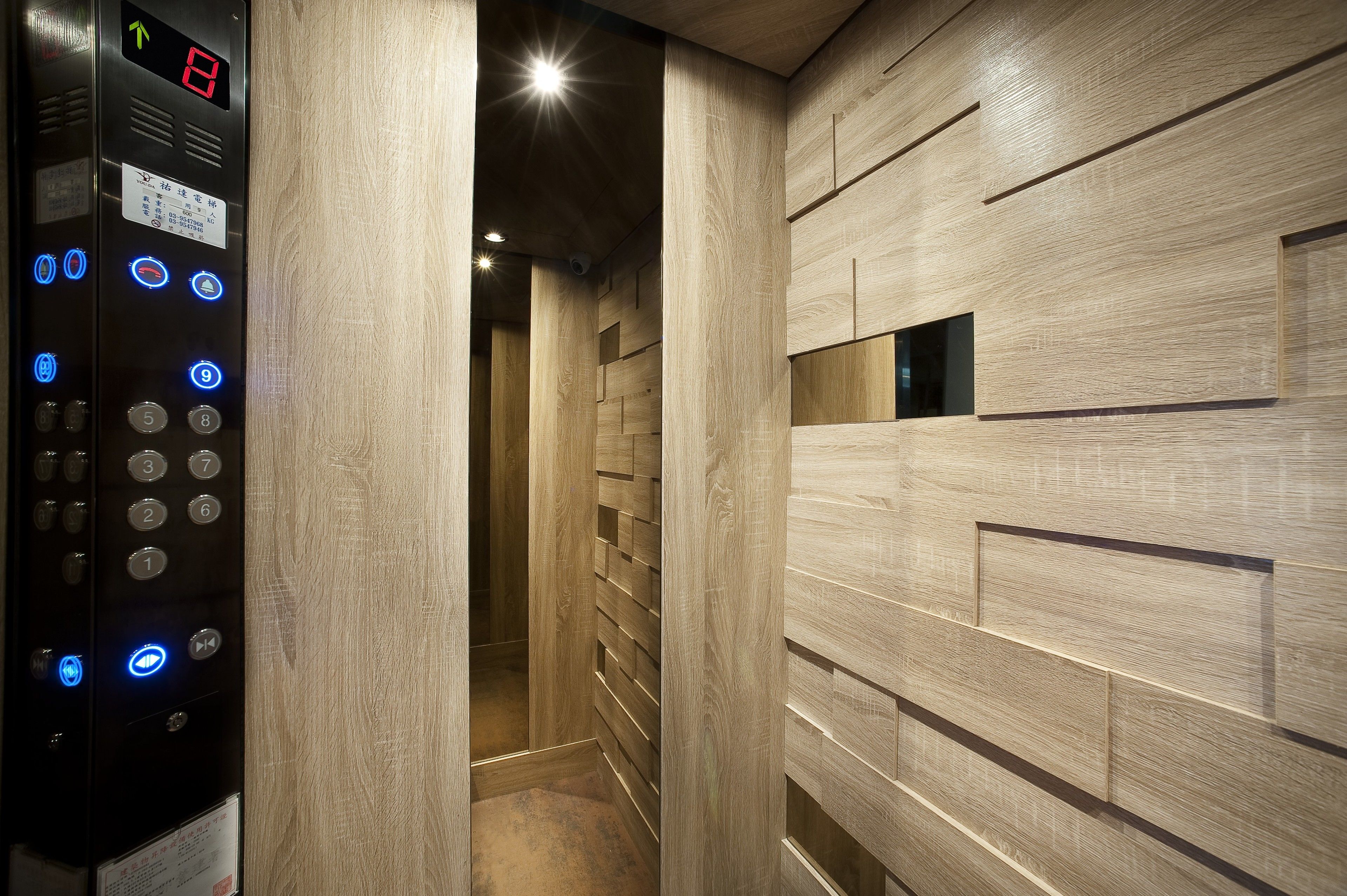 An elevator with a digital display and control panel, a wooden wall with a geometric pattern, and a corridor leading to another area.