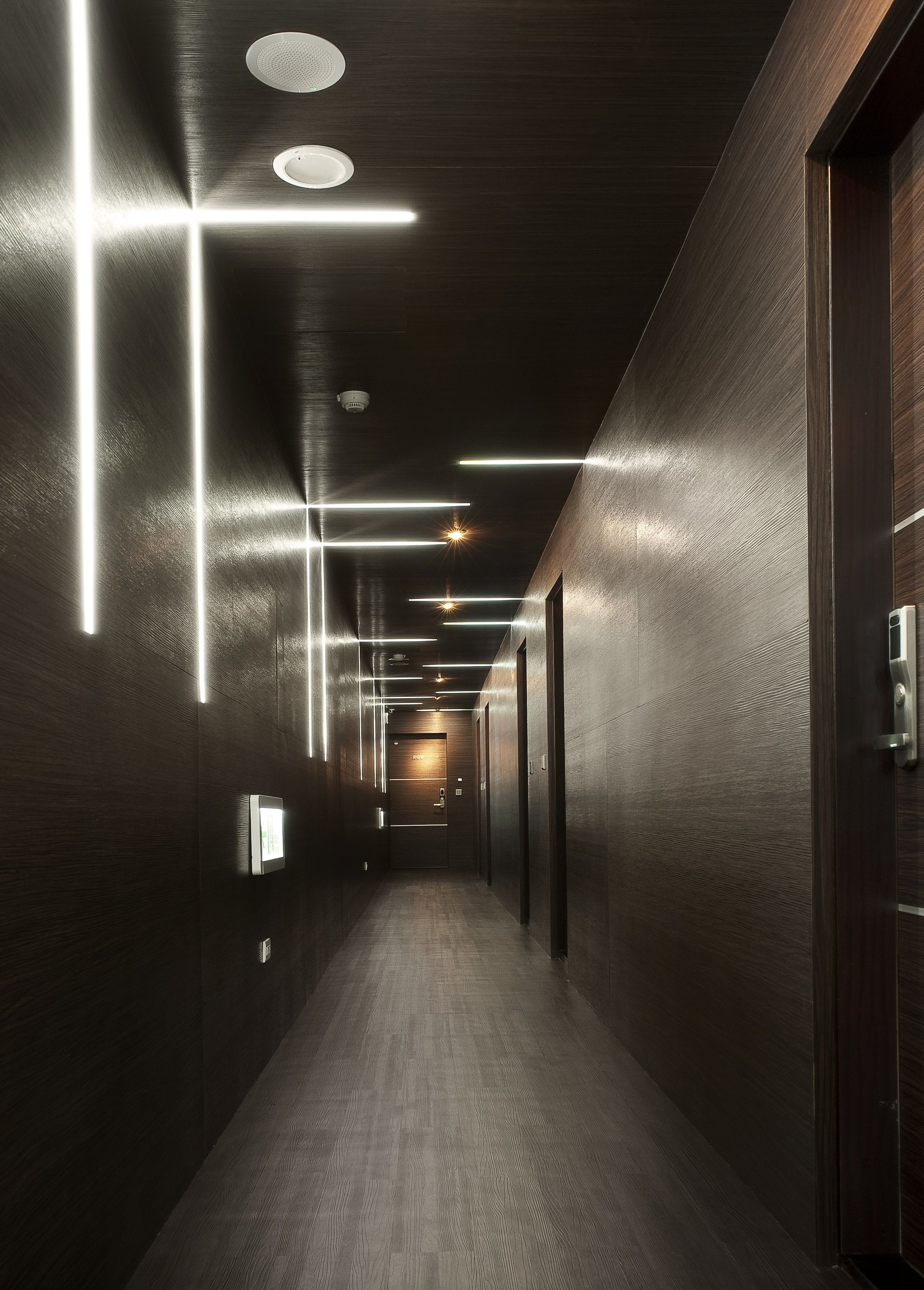 A modern hallway with a wooden floor and walls, featuring linear light fixtures and a security camera.