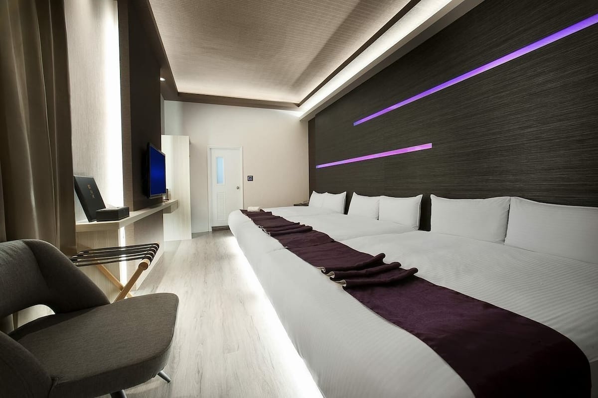 A modern hotel room with a long bed, a TV on a shelf, and a door in the background.