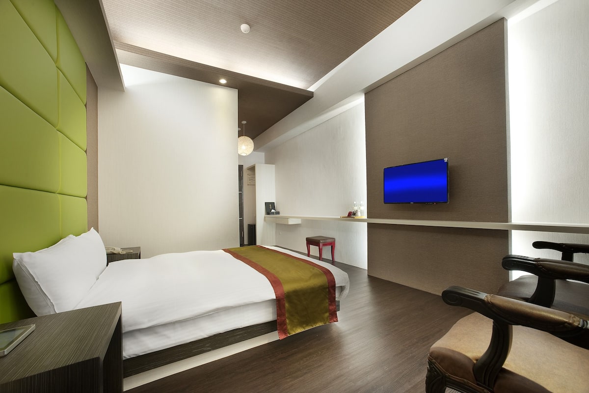 A modern hotel room with a bed, a desk, a chair, a TV, and a small kitchenette area.