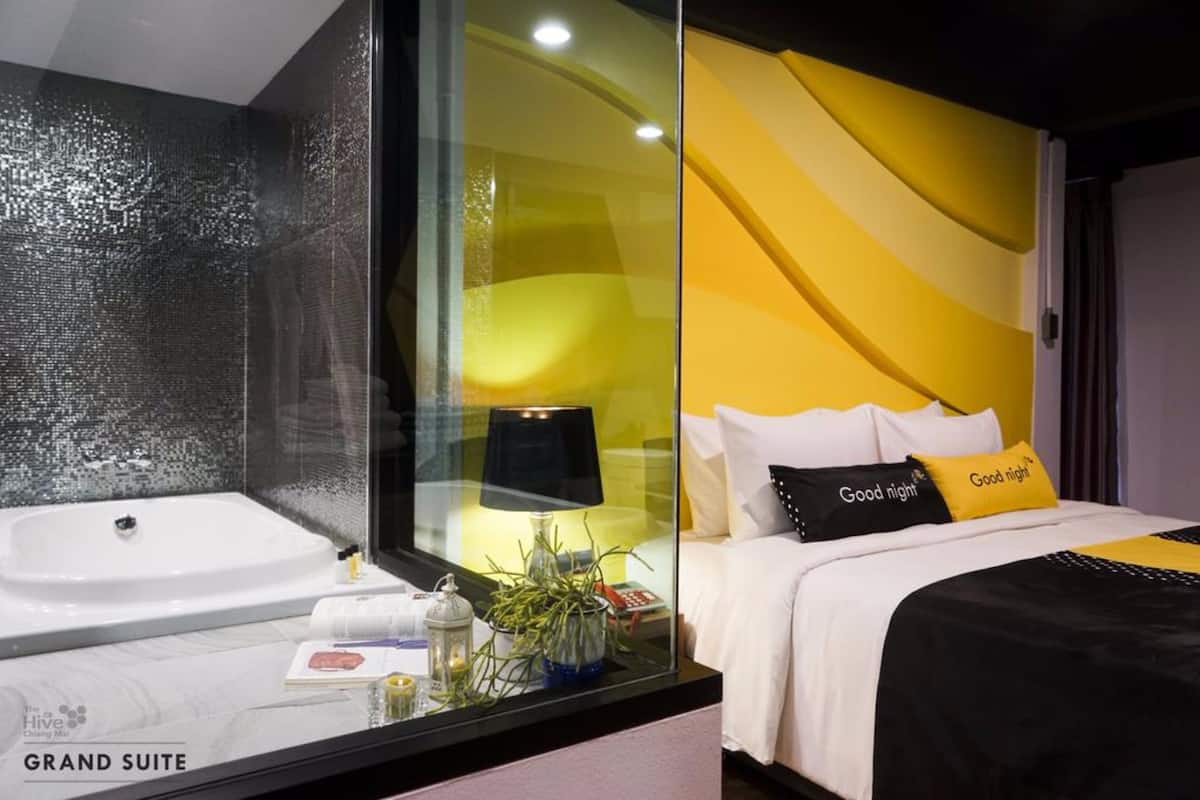 Una habitación de hotel moderna con un separador de vidrio, una cama grande con ropa de cama blanco y negro, y una mesita de noche pequeña con una lámpara y objetos decorativos.