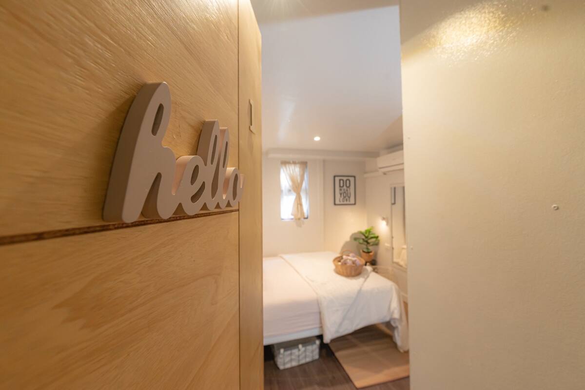 A wooden wall with a 'Hello' sign, a bed with a white bedspread, a small plant, and a framed picture on the wall.