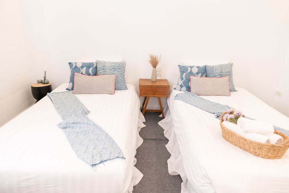 A room with two beds, each with a white bedspread and blue patterned pillows. There is a small wooden nightstand with a vase of dried flowers and a candle. A wicker basket with towels and a small vase of flowers is on the right bed.