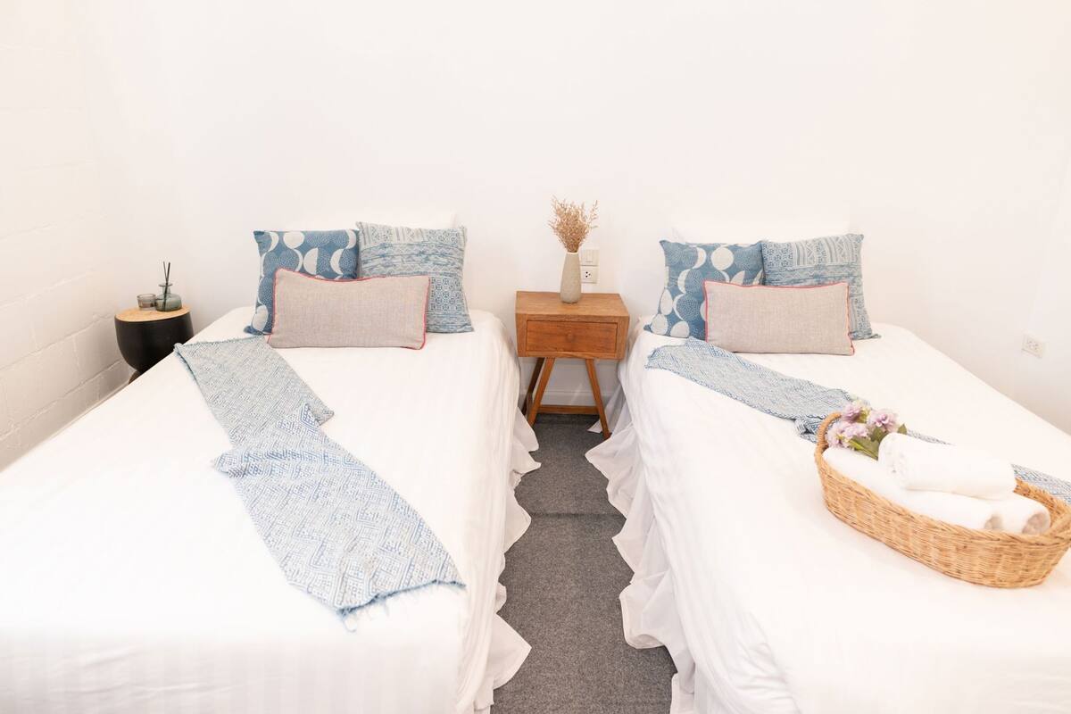A double room with two beds, each with a patterned pillow and a blue throw blanket. There is a nightstand with a vase and candles between the beds.
