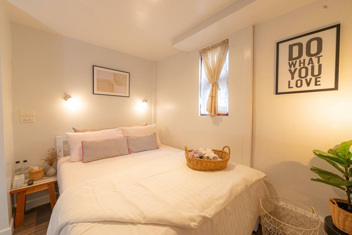 A neatly arranged bedroom with a bed, a bedside table, a wall-mounted light, a framed picture, a window with curtains, and a motivational poster.