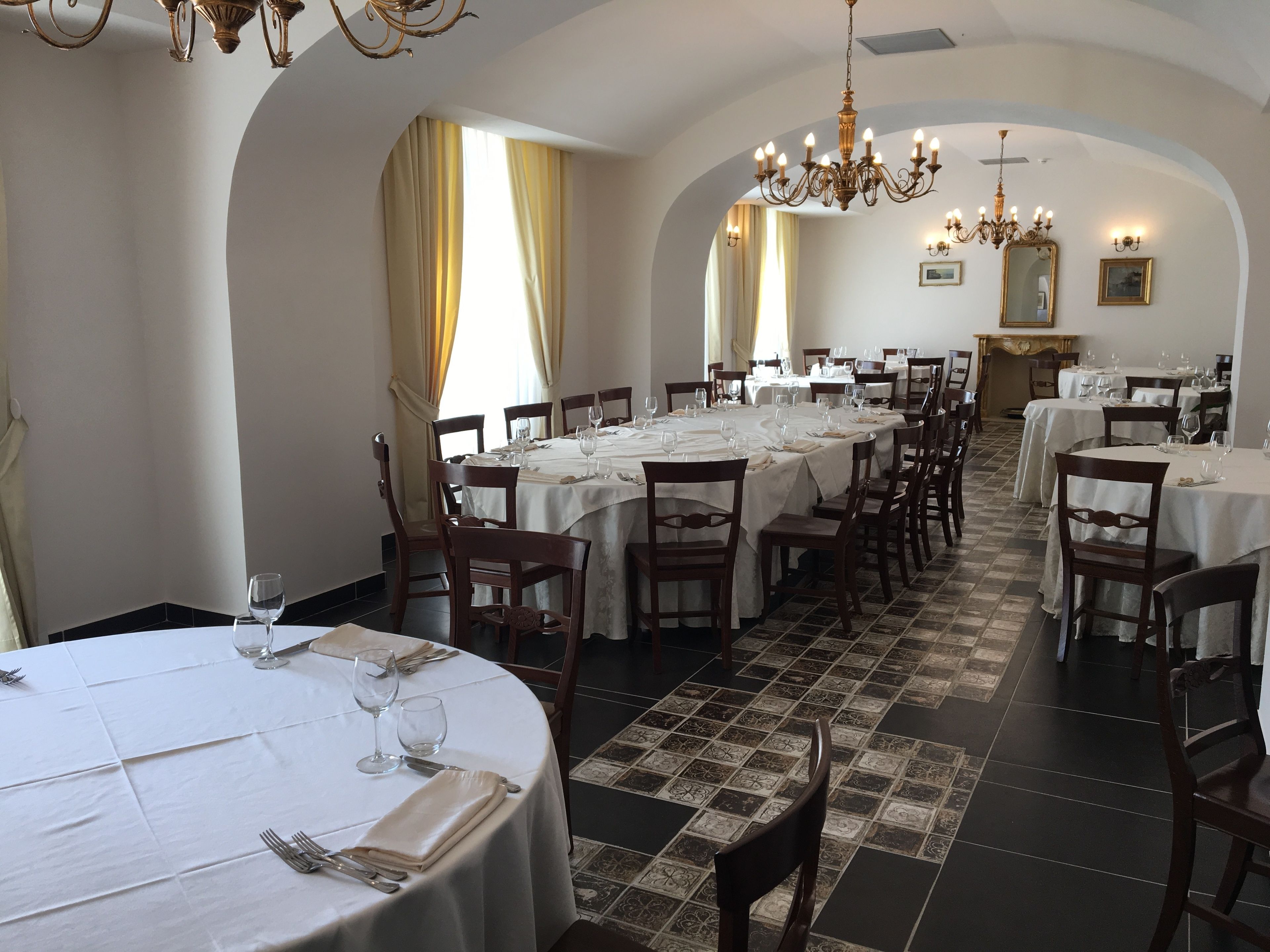 Un restaurante con varias mesas redondas, cada una con manteles blancos, copas de vino y cubiertos. El espacio cuenta con techos abovedados, candelabros y baldosas de suelo con diseño.
