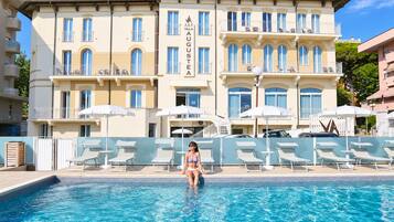 Area piscina con lettini e ombrelloni, un edificio con balconi e un cartello con la scritta 'VILLA AUGUSTEA'.