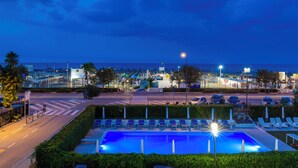 A nighttime view of a pool area with lounge chairs, a road with cars, and a distant cityscape. - Villa Augustea (Rimini)