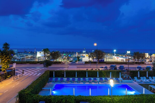 A nighttime view of a pool area with lounge chairs, a road with cars, and a distant cityscape. - Villa Augustea (Rimini)