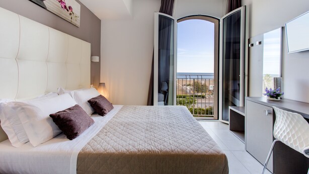 A hotel room with a bed, a desk, and a view of the city through a balcony. - Villa Augustea (Rimini)