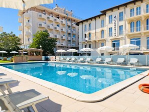 A swimming pool with lounge chairs and umbrellas, surrounded by multi-story buildings with balconies. - Villa Augustea (Rimini)