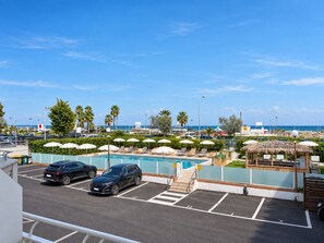 A pool area with lounge chairs and umbrellas, a parking lot with two cars, and a view of the sea. - Villa Augustea (Rimini)