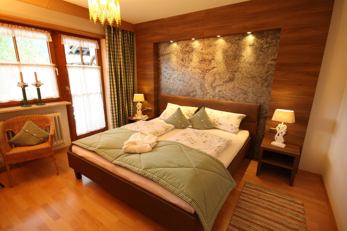 Un dormitorio con cabecera de madera, una cama grande con ropa de cama verde, dos mesitas de noche con lámparas, una silla y un ventanal con cortinas.