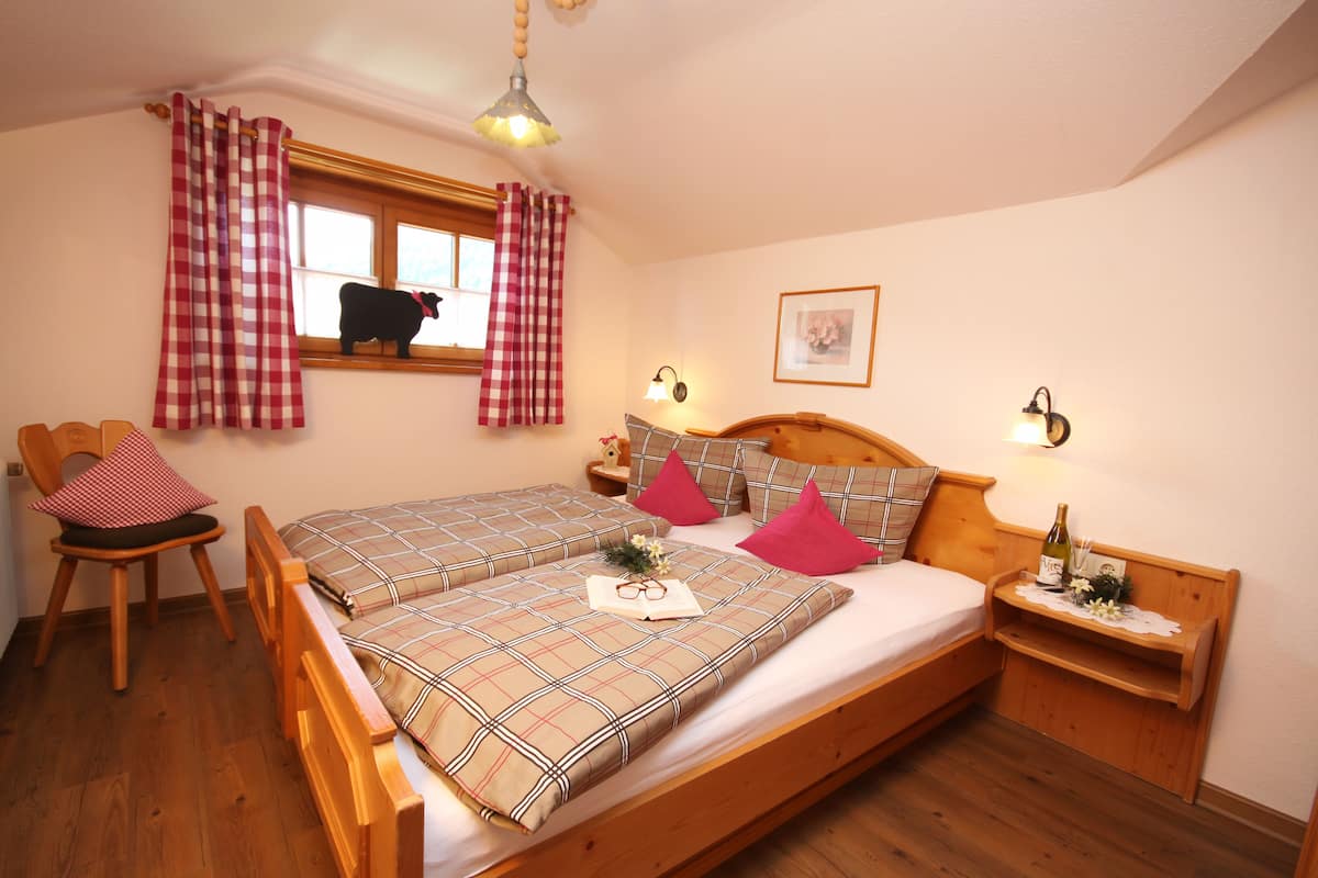 Un dormitorio con cama de madera, cortinas a cuadros, una ventana con un gato negro, una silla de madera y una mesita de noche con una botella.