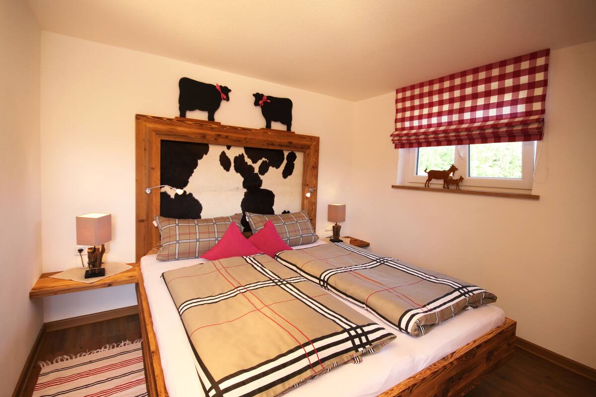 Un dormitorio con cabecera de madera, dos mesitas de noche con lámparas, una ventana con persiana a cuadros y una pequeña figura decorativa de vaca en el alféizar.