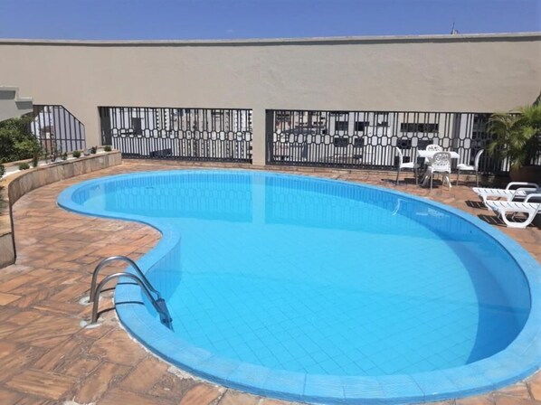Una piscina con borde curvo, rodeada por una terraza con baldosas. Se observan sillones de descanso y una pequeña mesa al fondo.