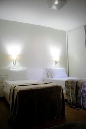Habitación de hotel con dos camas, cada una con cabecero blanco y una lámpara de noche. El piso es de madera y hay una ventana a la derecha.