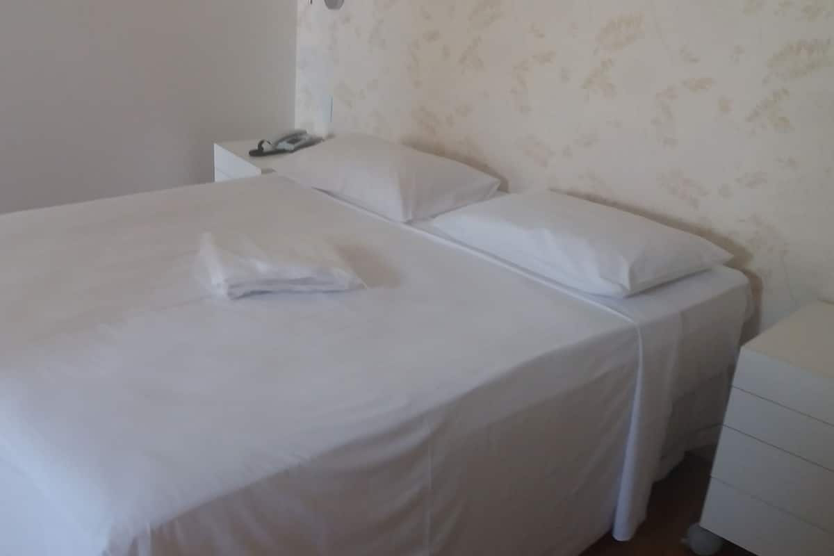 A neatly made bed with white linens and pillows, a bedside table with a lamp, and a patterned wallpaper on the wall.