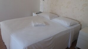 Una cama bien hecha con sábanas y almohadas blancas, una mesita de noche con una lámpara y un papel tapiz estampado en la pared.