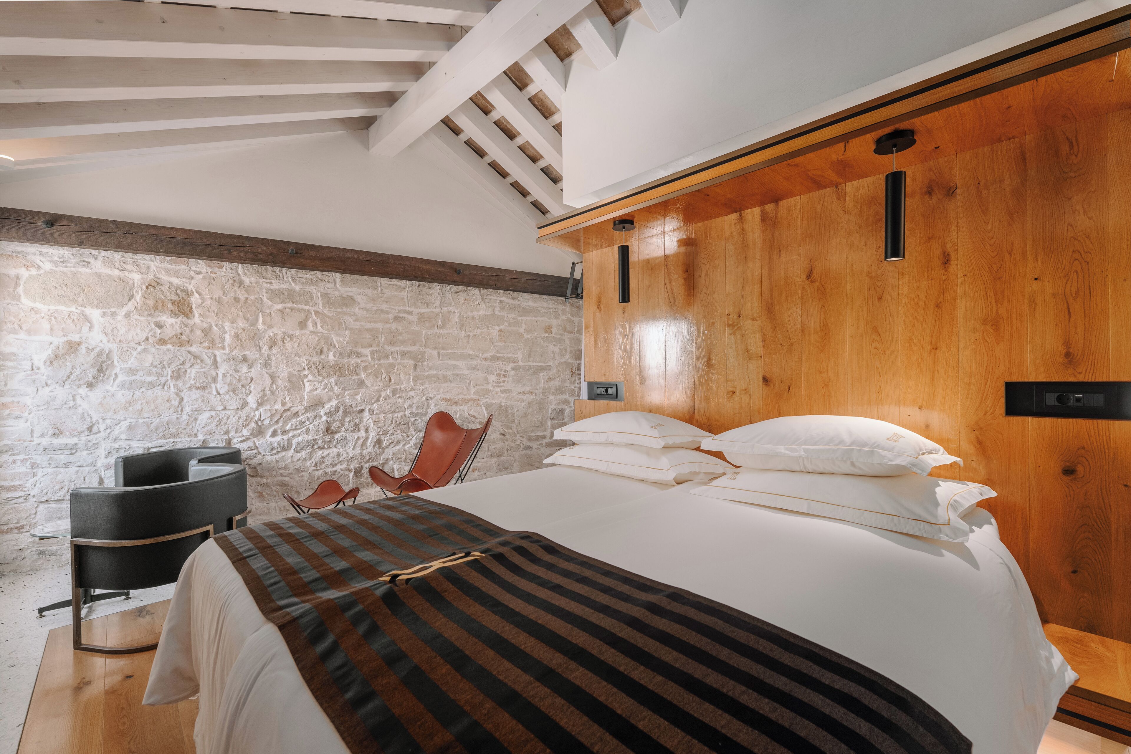 Ein Schlafzimmer mit einem großen Bett, einem Holzkopfende, einer Steinwand und einem modernen Sessel.
