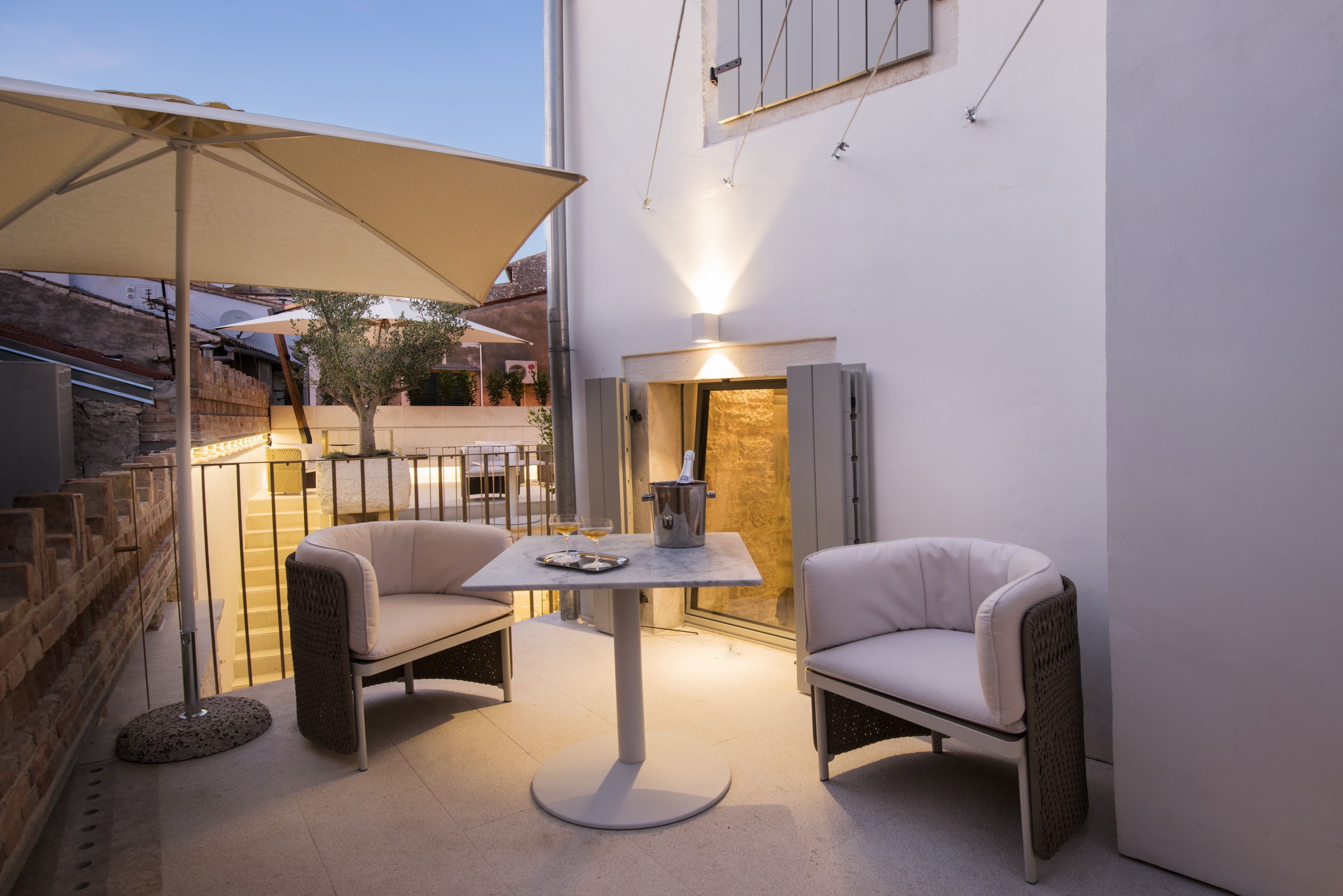 Un área de estar al aire libre con dos sillones acolchados y una mesa, un paraguas grande y una escalera que lleva a un nivel superior.