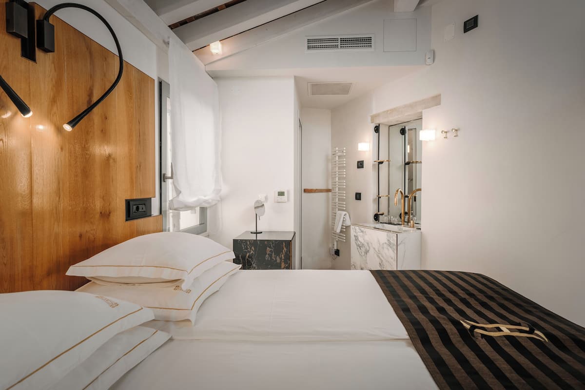 Habitación de hotel moderna con cabecera de madera, cama con ropa de cama blanca, mesita de noche con lámpara y baño con espejo y perchero para toallas.