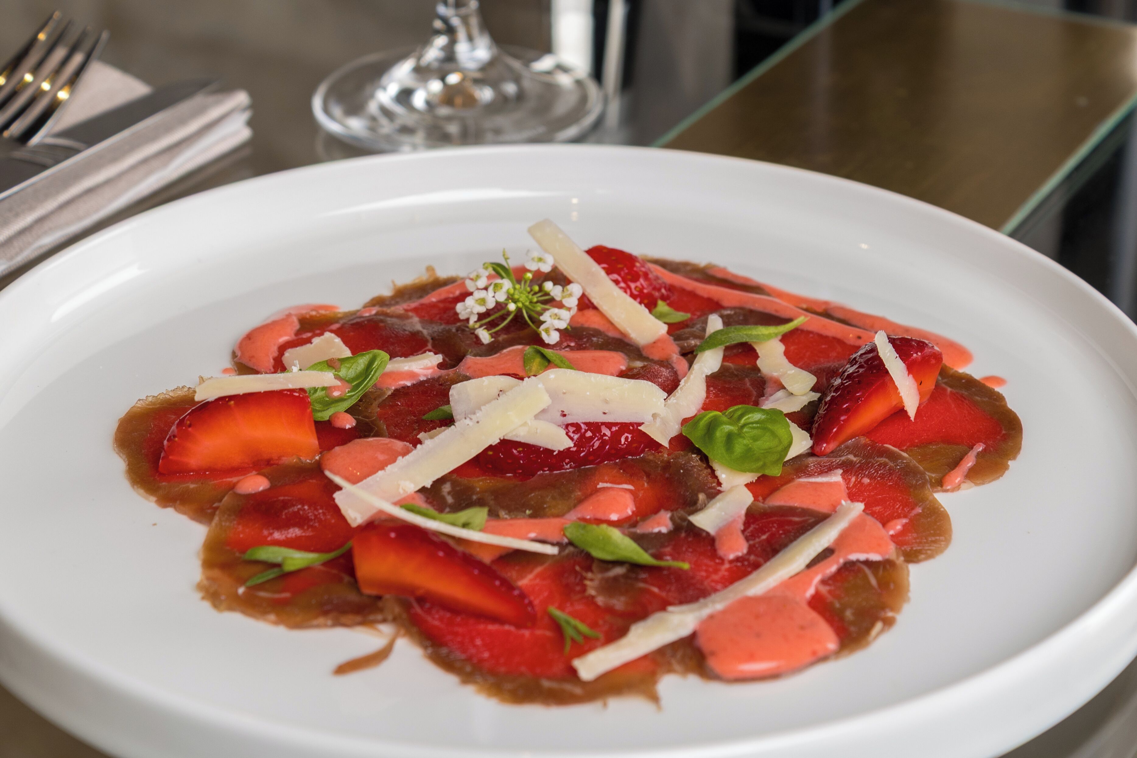 A white plate with a dish of sliced strawberries and a drizzle of sauce, garnished with cheese and basil.