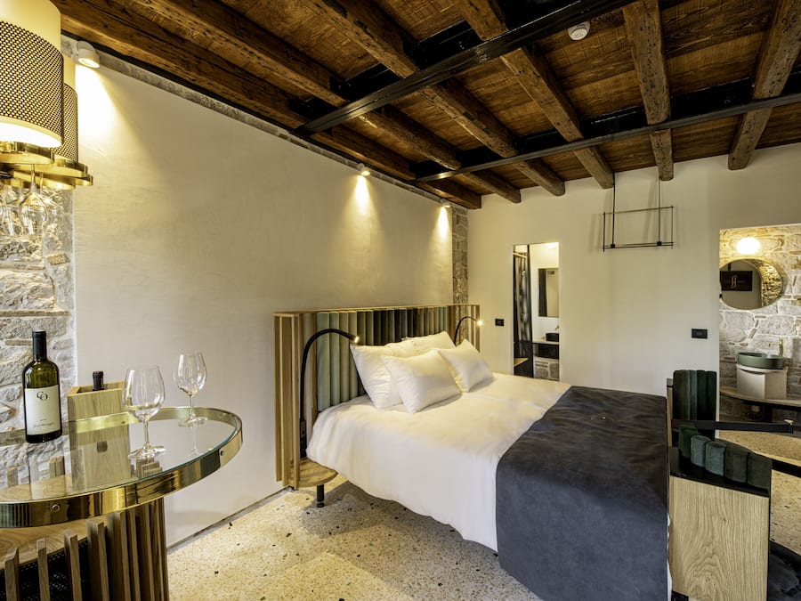 Ein Schlafzimmer mit einem Bett, einem Nachttisch mit einer Weinflasche und Gläsern, einer Steinsäule und einer Holzbalkendecke.