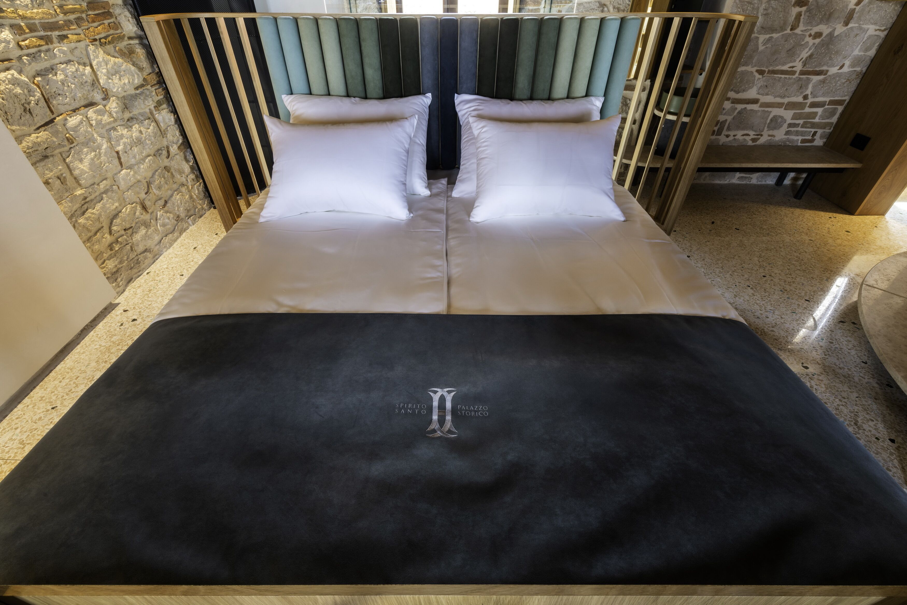 A bed with a black cover, a stone wall, and a wooden headboard.