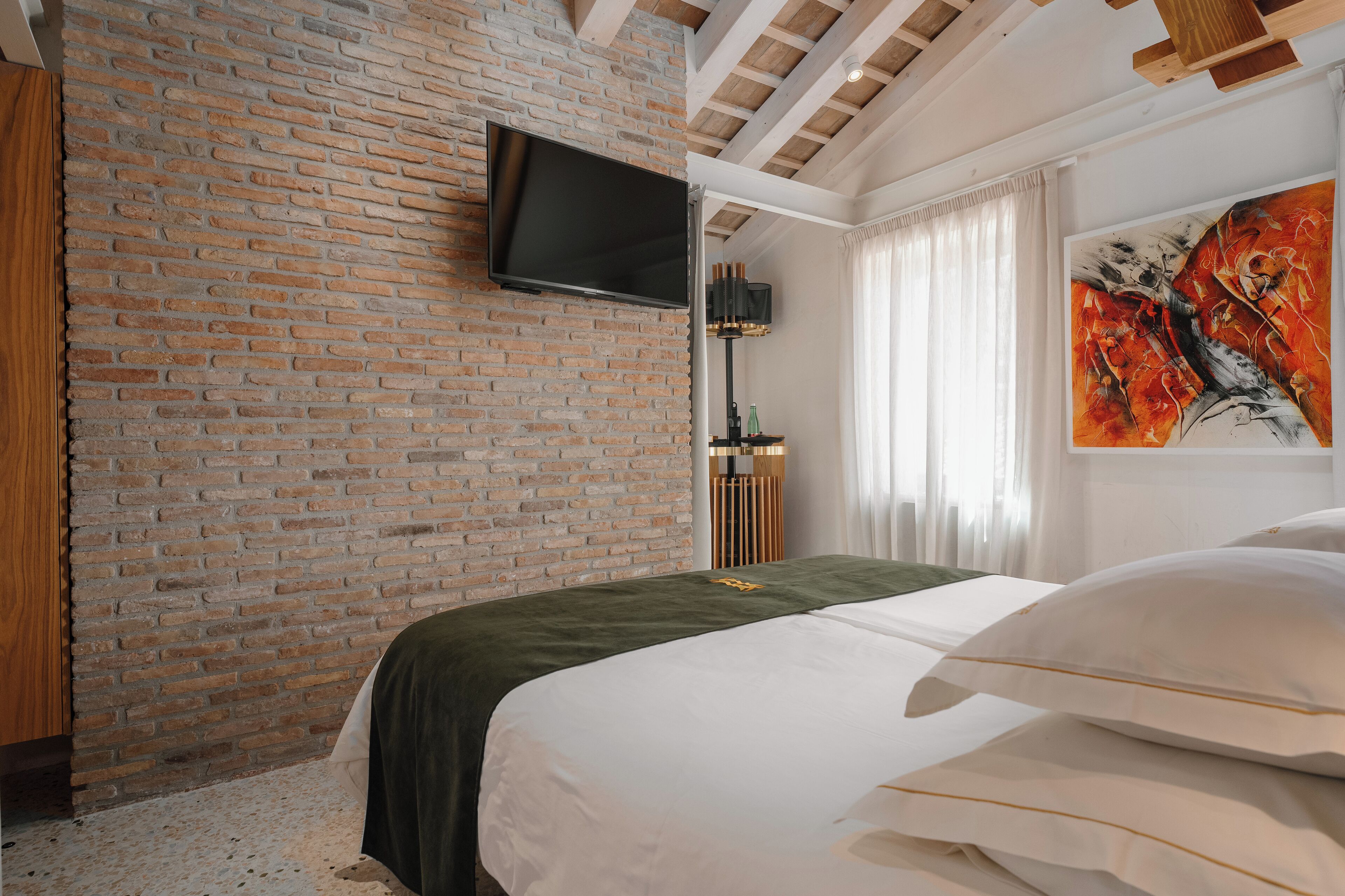 A bedroom with a brick wall, a flat-screen TV mounted above the bed, a large abstract painting, and a green bedspread.