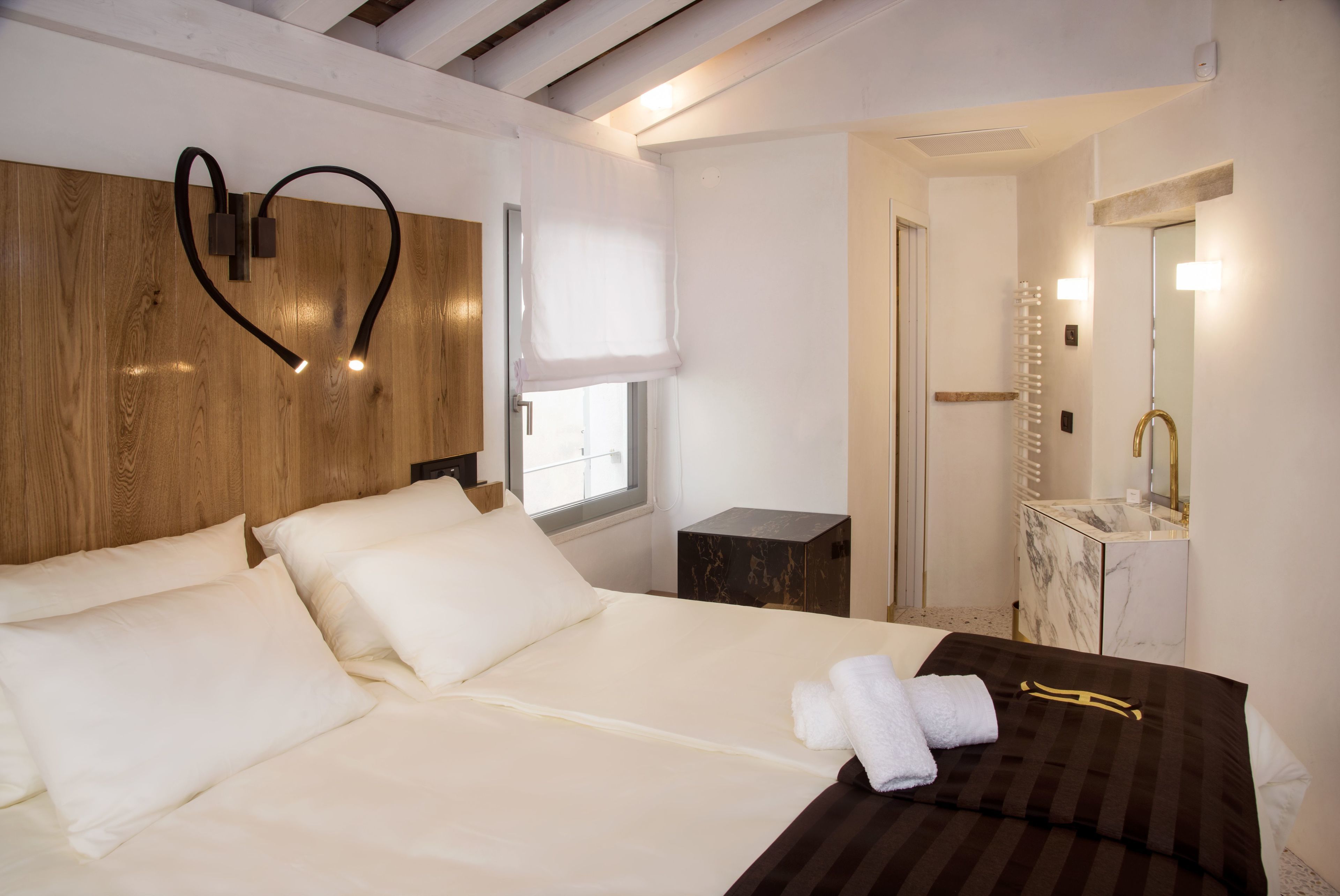 A hotel room with a wooden headboard, a large bed with white linens, a bedside table with a lamp, and a bathroom with a marble vanity and mirror.