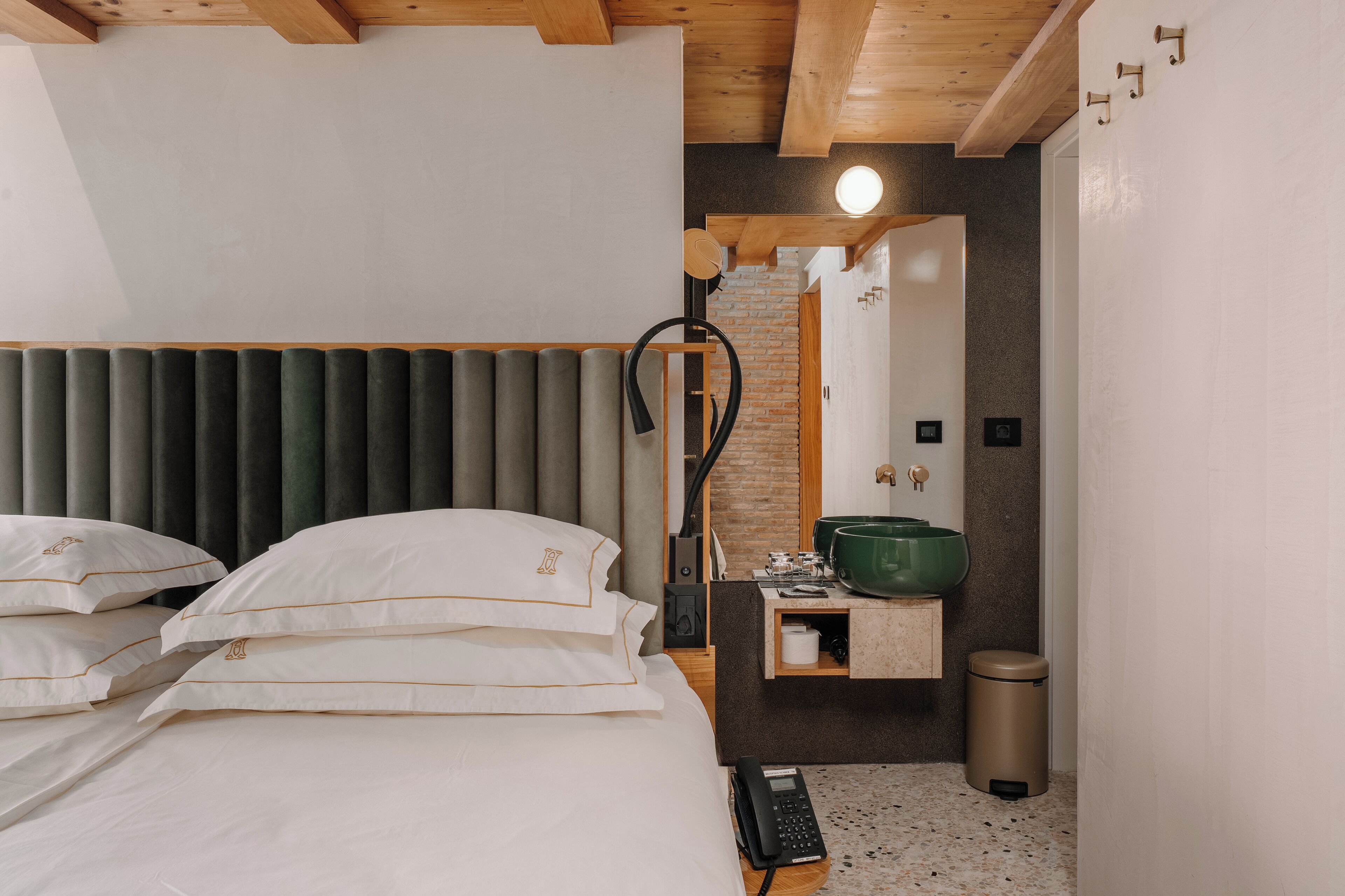 A hotel room with a bed, a bedside table with a green bowl, a trash bin, and a mirror.