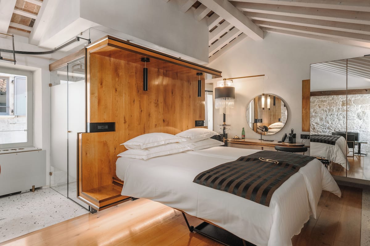 Un dormitorio moderno con cabecera de madera, una cama grande, mesitas de noche, un espejo y una pared de piedra.
