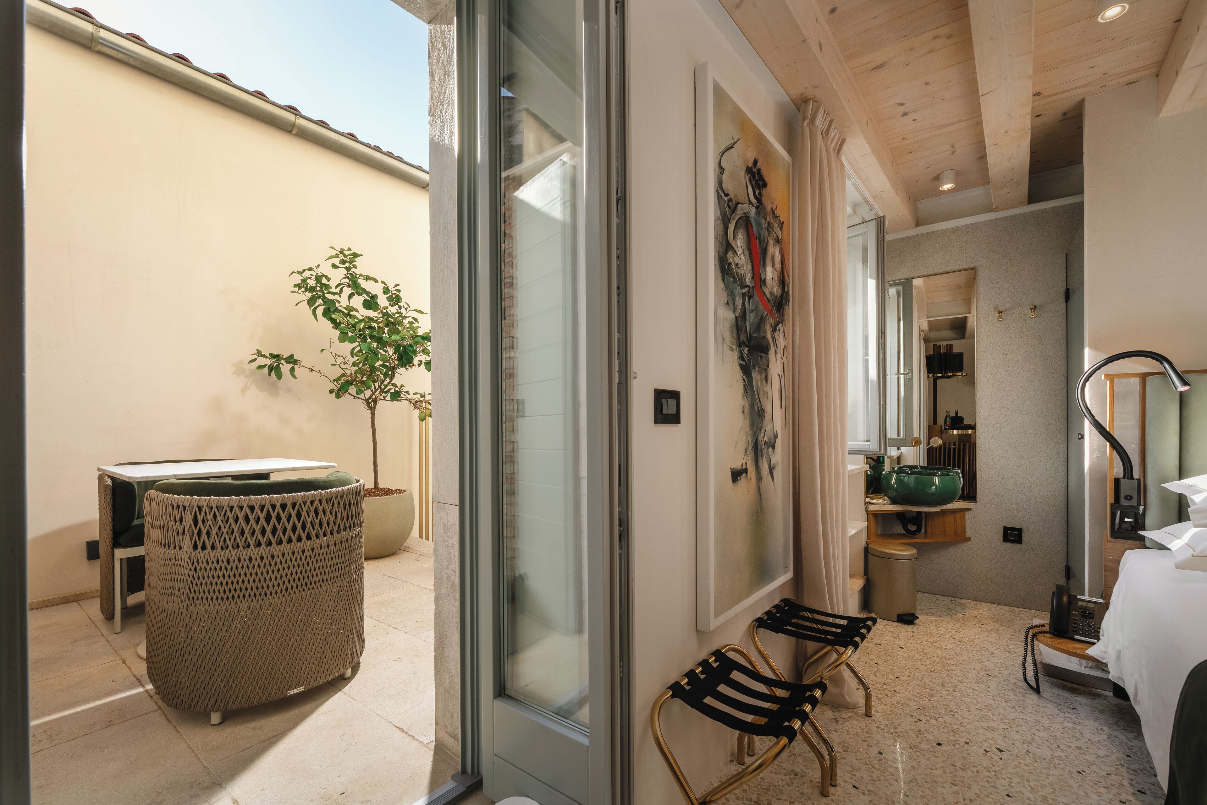 A hotel room with a large window, a wicker chair, a small table, a green plant, and a bathroom with a sink and mirror.