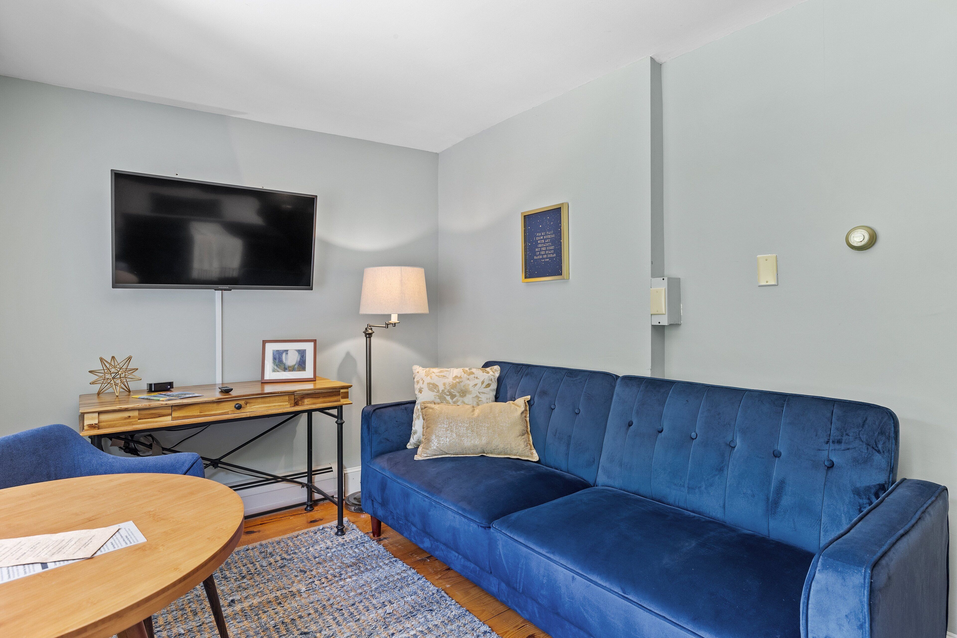 Uma sala de estar moderna com um sofá azul, uma escrivaninha de madeira, uma TV de tela plana e um abajur.