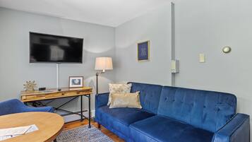 Uma sala de estar moderna com um sofá azul, uma escrivaninha de madeira, uma TV de tela plana e um abajur.