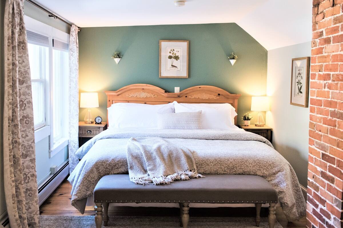 A bedroom with a wooden bed, two bedside tables with lamps, a bench, and a framed botanical print on the wall.