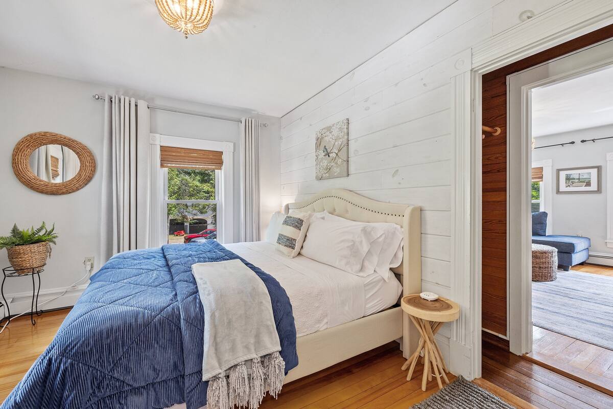A bedroom with a bed, a bedside table, a wall-mounted mirror, a window with curtains, and a view of the outside.