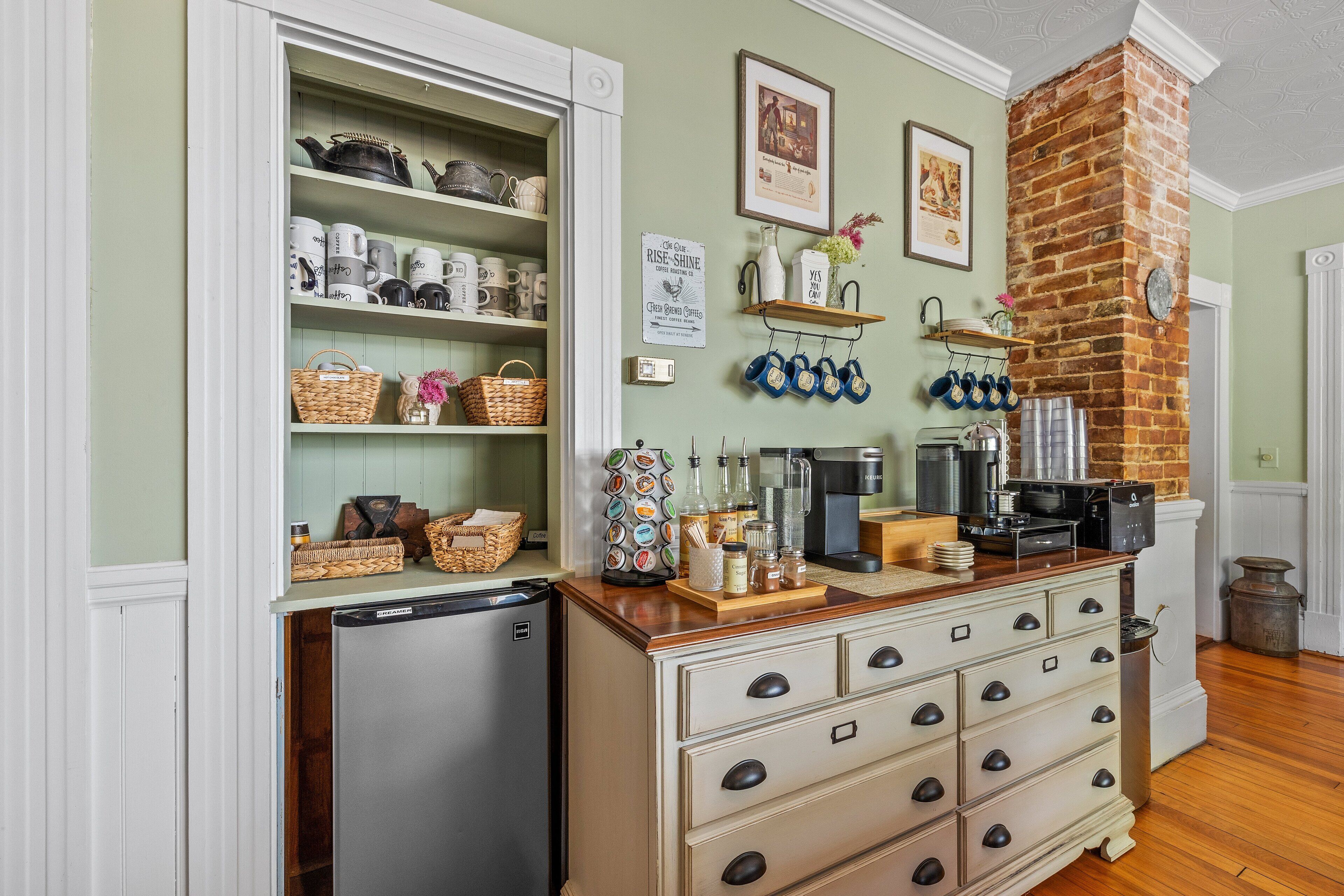 Una cocina con refrigerador, un mueble de madera con cajones, estantes con utensilios de cocina, una pared de ladrillo y una máquina de café.