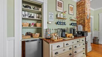 Una cocina con refrigerador, un mueble de madera con cajones, estantes con utensilios de cocina, una pared de ladrillo y una máquina de café.