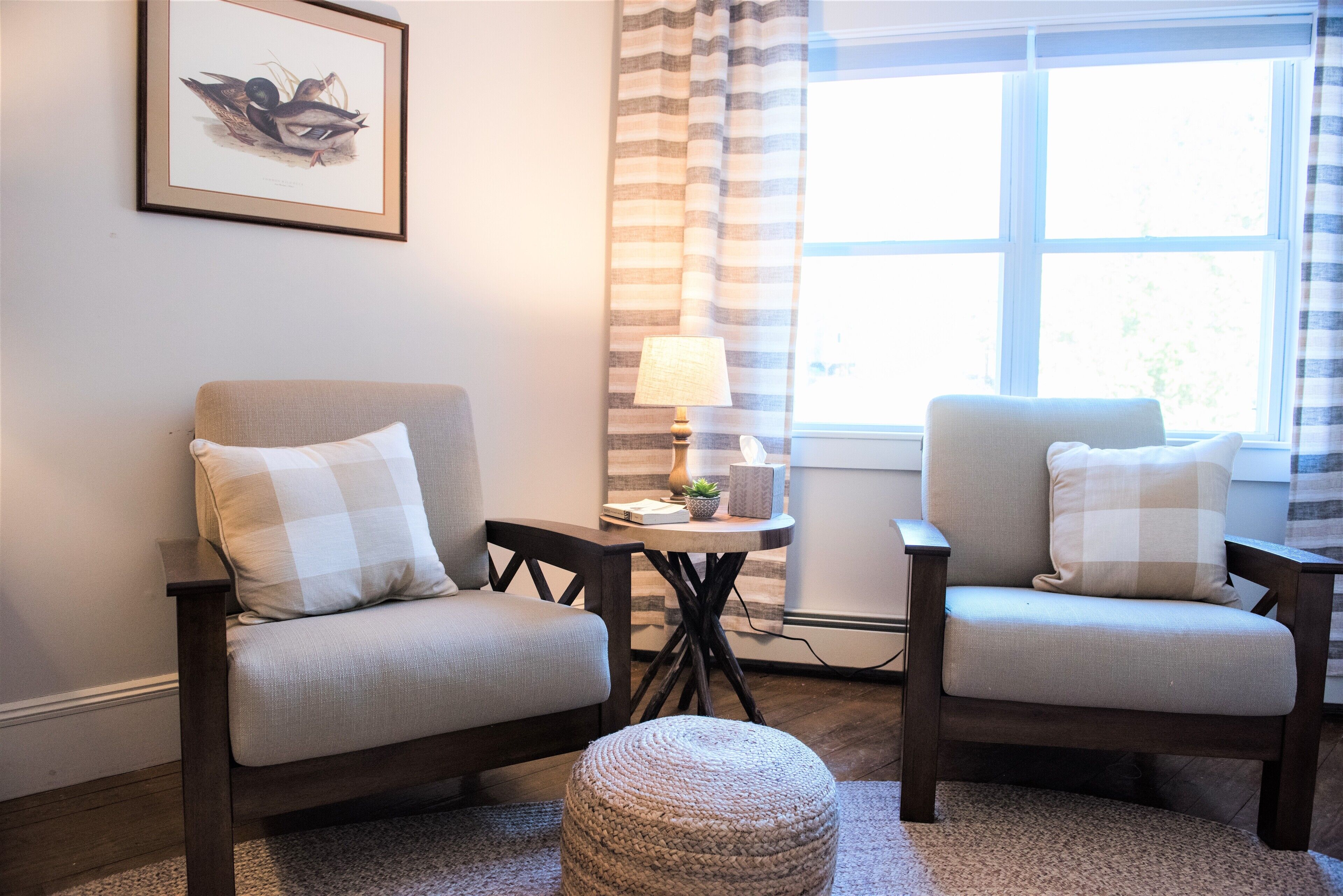 A room with two armchairs, a small table with a lamp, a framed picture on the wall, and a window with curtains.