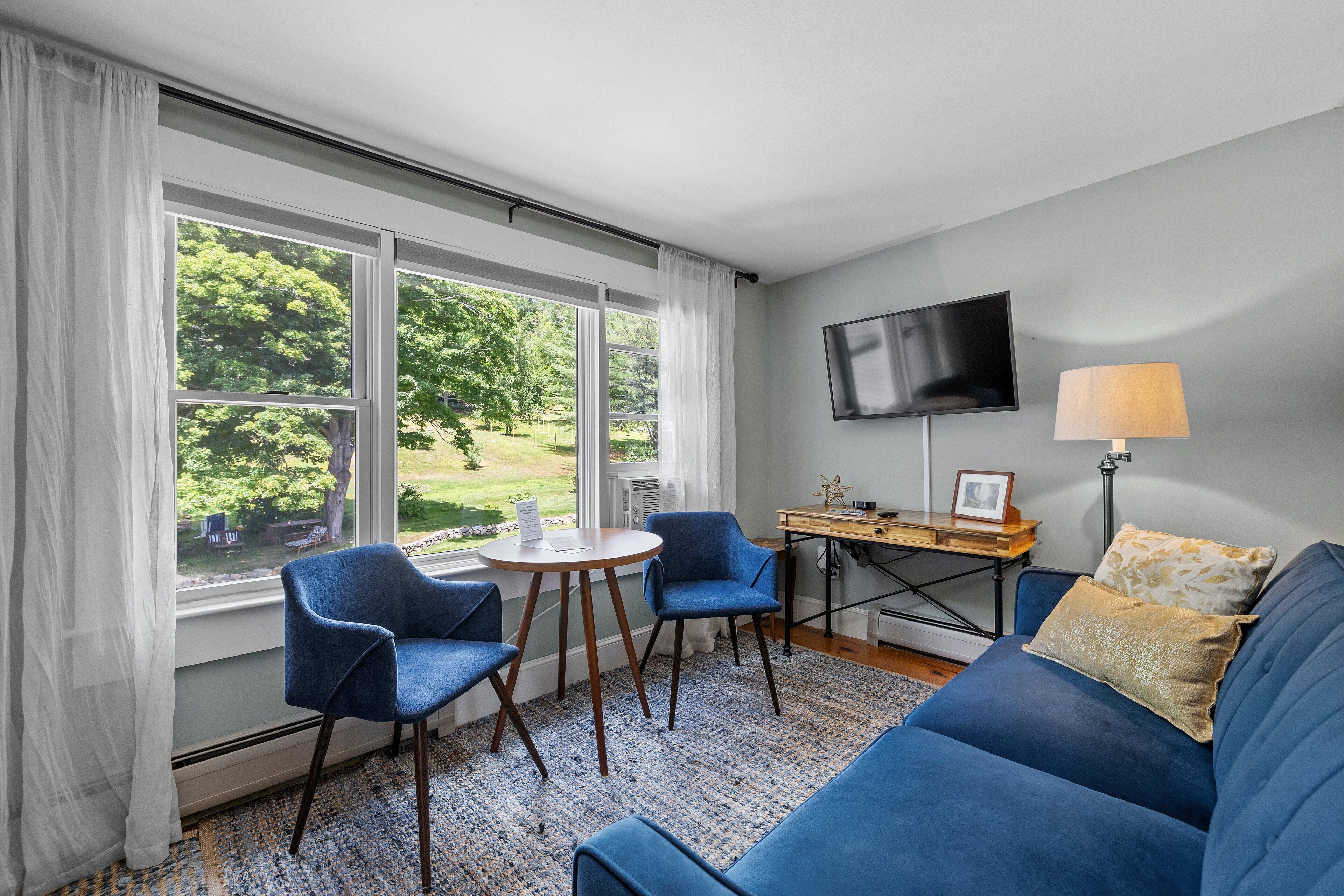 Une pièce avec un canapé bleu, deux fauteuils bleus, une petite table ronde, un téléviseur fixé au mur et une fenêtre avec des rideaux.