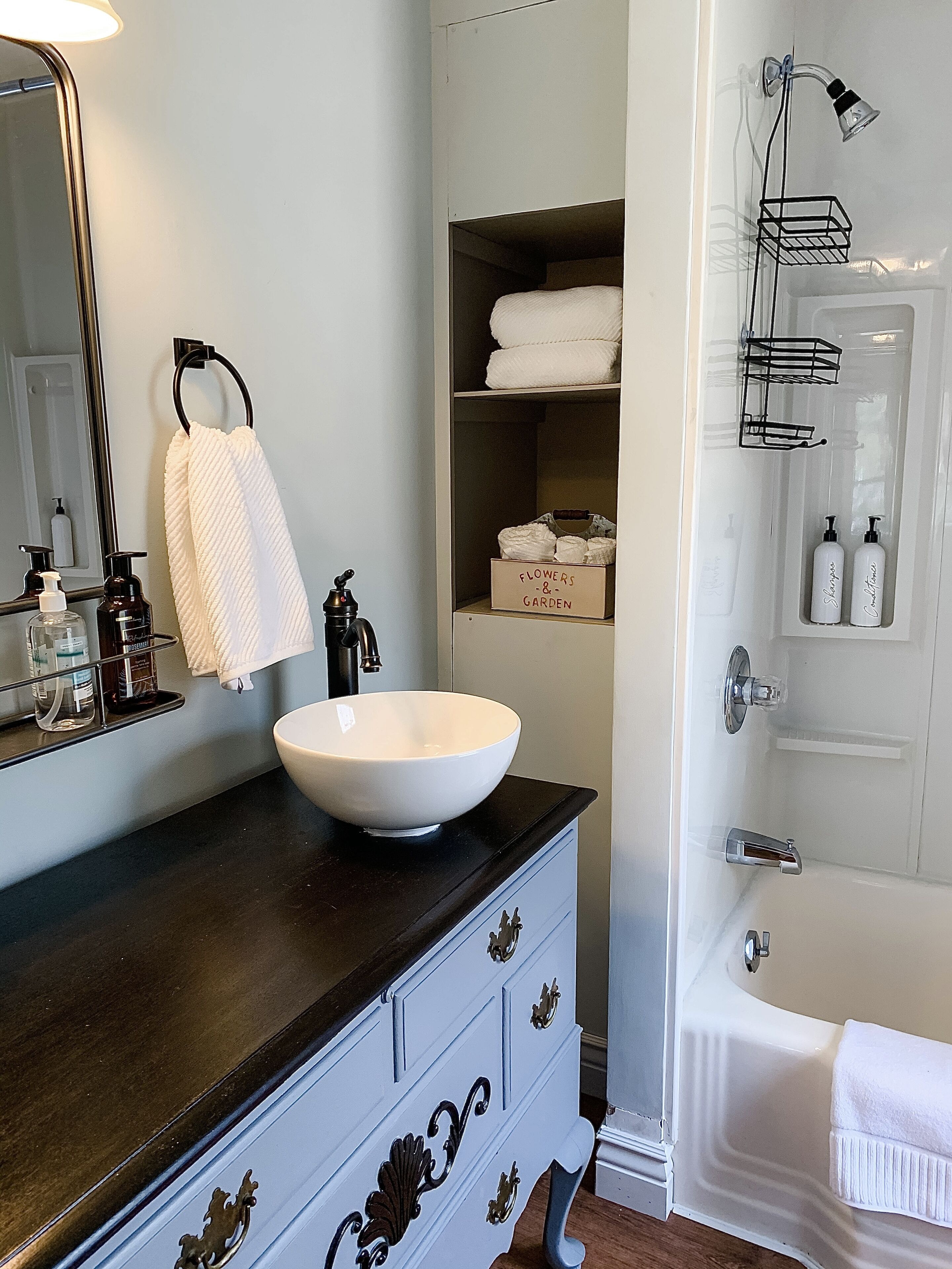 Une salle de bain avec un lavabo blanc, un plan de toilette foncé et un miroir. On y trouve des articles de toilette sur le plan de toilette et une serviette accrochée au mur.