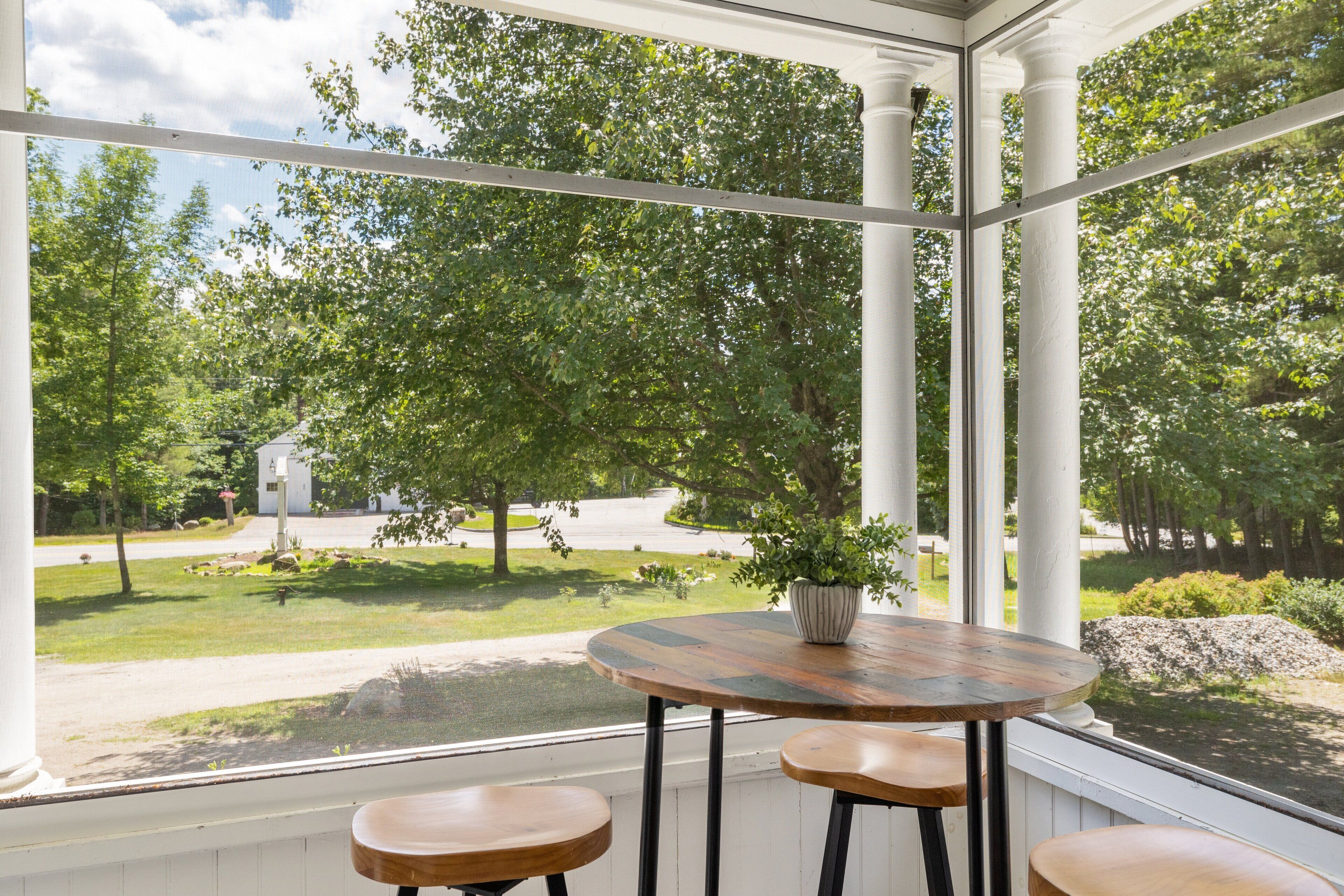 A conservatory with a round wooden table and three stools, a potted plant, and a view of a green landscape with trees and a white building.