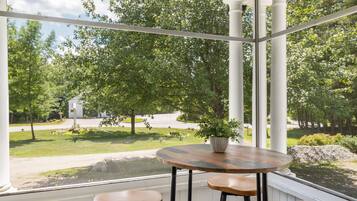 A conservatory with a round wooden table and three stools, a potted plant, and a view of a green landscape with trees and a white building.