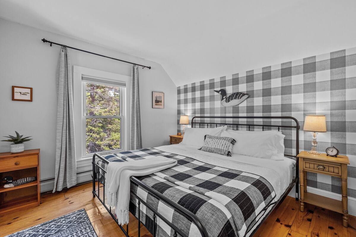 A bedroom with a checkered headboard, a window with curtains, a bedside table with a lamp, and a nightstand with a clock.