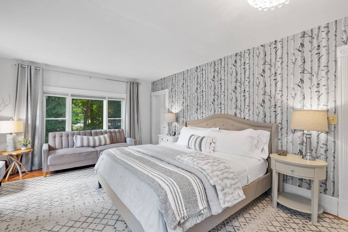 A bedroom with a large bed, a sofa, two bedside tables with lamps, and a window with curtains.