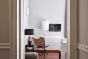 Una camera moderna con una TV a schermo piatto montata a parete, una lampada da terra, una sedia di legno e un piccolo tavolino con lampada.
