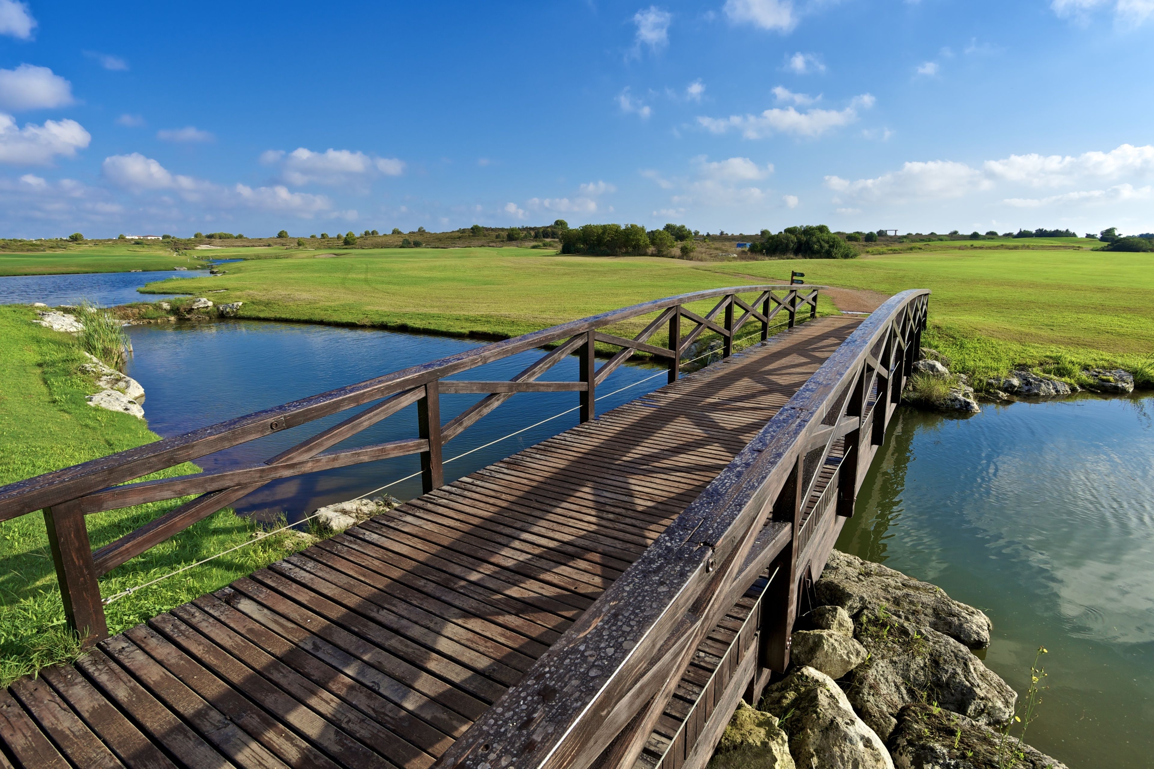 A wooden bridge over a calm river with grassy banks and a clear blue sky.