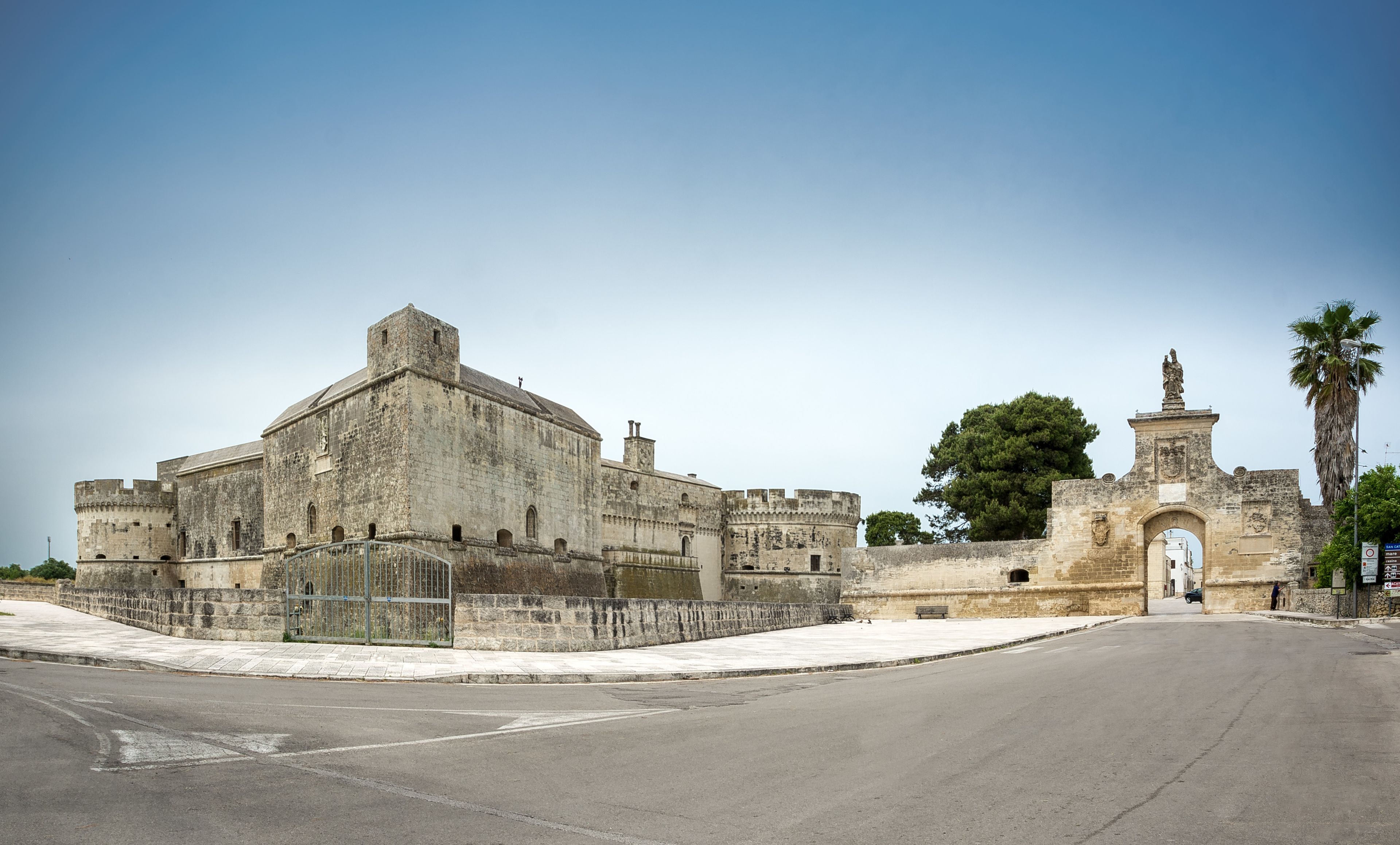 A historic stone building with a large central tower, a fortified entrance, and a palm tree in the background.